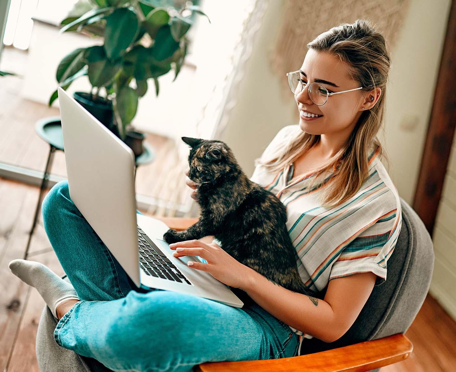 A woman with glasses smiles while working on a laptop with her cat